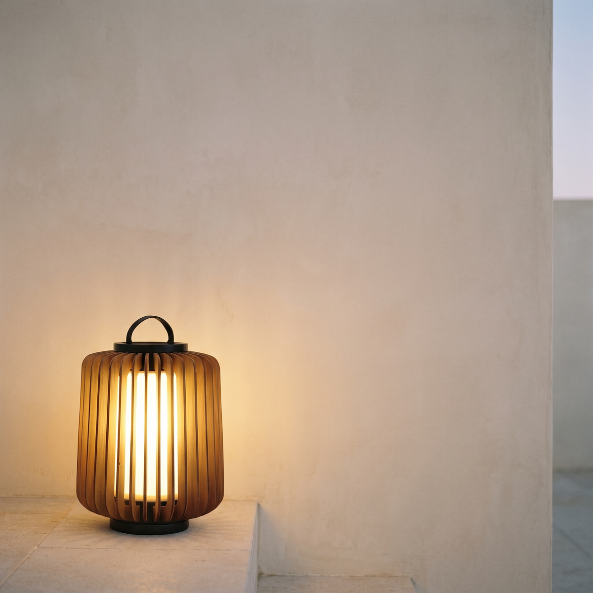Slatted teak outdoor lantern glowing on a limestone step against a bone plaster garden wall at dusk